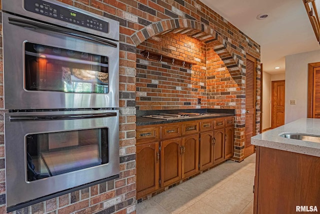 kitchen with stainless steel appliances, brown cabinetry, light tile patterned flooring, dark stone countertops, and brick wall