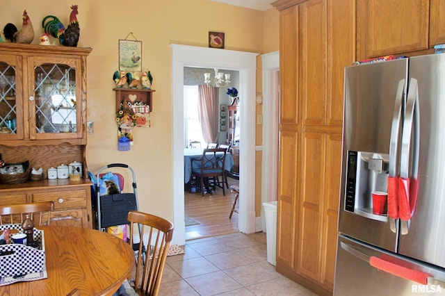 kitchen featuring light tile patterned floors, stainless steel refrigerator with ice dispenser, brown cabinetry, glass insert cabinets, and an inviting chandelier