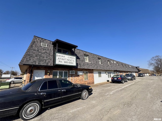 view of front of house featuring uncovered parking, roof with shingles, brick siding, and mansard roof