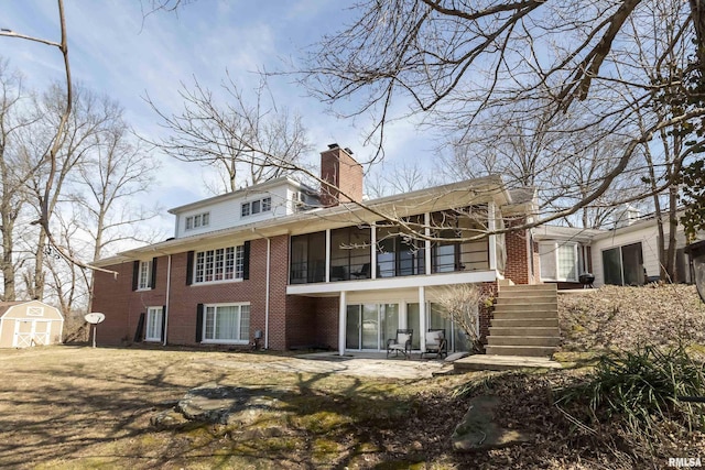 back of house with brick siding, stairs, a chimney, a storage shed, and an outbuilding