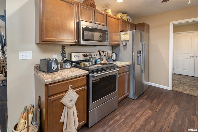 kitchen with dark wood-type flooring, baseboards, light stone counters, brown cabinetry, and stainless steel appliances