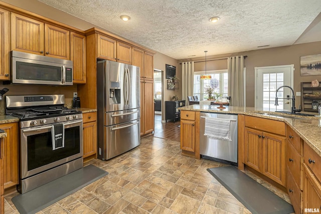 kitchen featuring hanging light fixtures, light stone counters, brown cabinetry, stainless steel appliances, and a sink