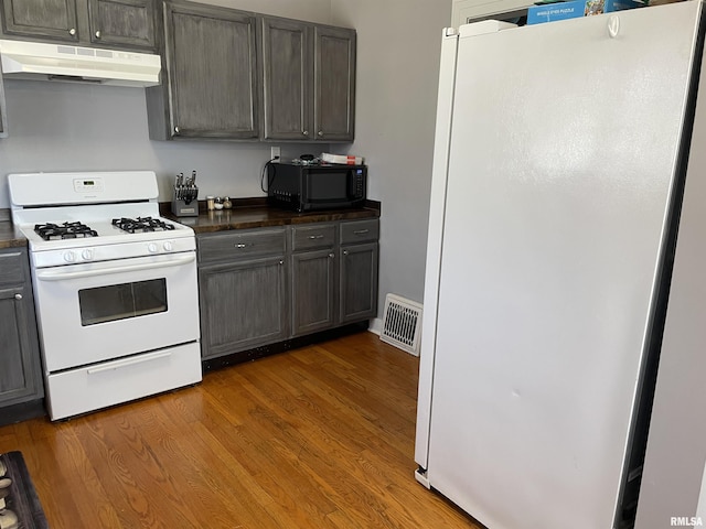 kitchen featuring dark countertops, visible vents, under cabinet range hood, wood finished floors, and white appliances