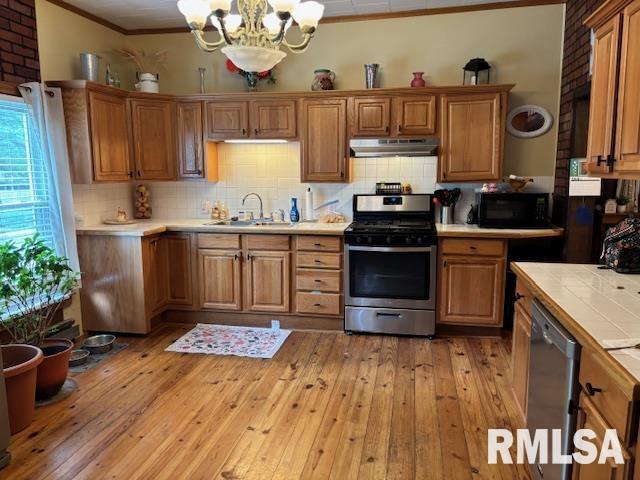 kitchen with appliances with stainless steel finishes, an inviting chandelier, a sink, tile countertops, and under cabinet range hood
