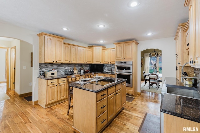 kitchen with light brown cabinetry, black electric stovetop, stainless steel double oven, arched walkways, and backsplash