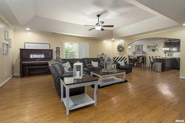 living area featuring ornamental molding, ceiling fan, arched walkways, a tray ceiling, and light wood-style floors