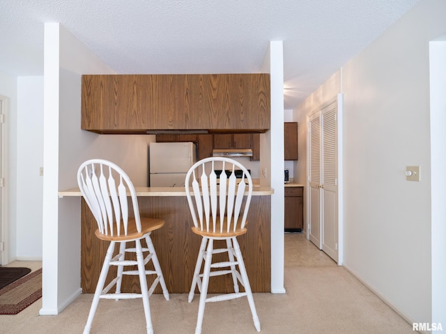 kitchen featuring a textured ceiling, under cabinet range hood, light carpet, freestanding refrigerator, and brown cabinets