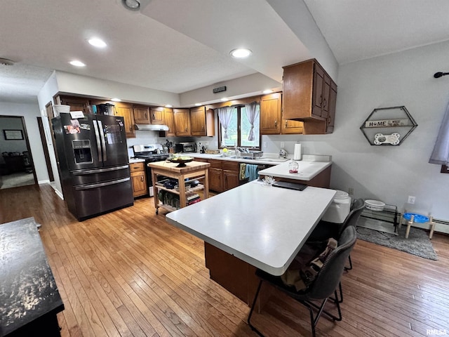 kitchen featuring light wood finished floors, under cabinet range hood, stainless steel range with gas stovetop, a breakfast bar, and black fridge