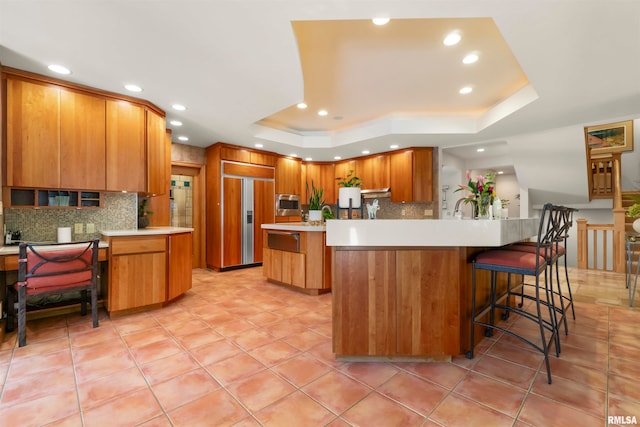 kitchen featuring a breakfast bar, a tray ceiling, a center island, built in appliances, and decorative backsplash