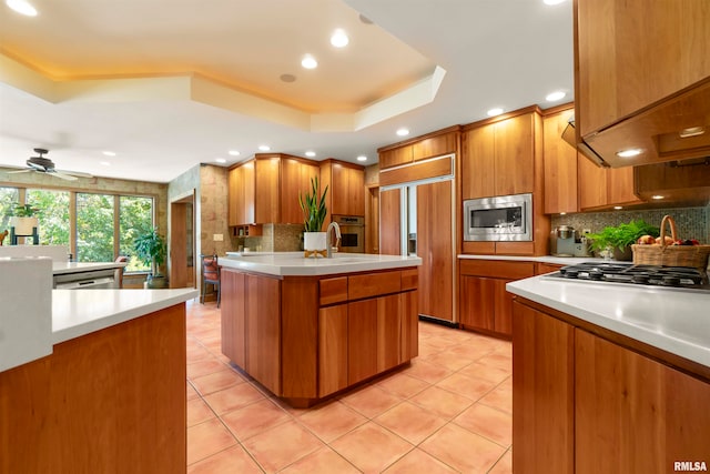 kitchen featuring a raised ceiling, backsplash, a kitchen island, built in appliances, and ceiling fan