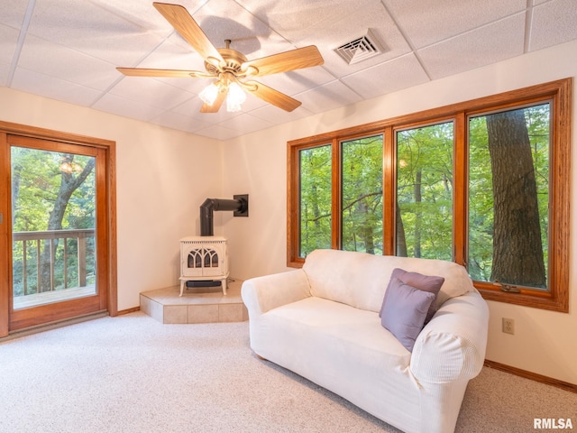 carpeted living room with a drop ceiling, a wood stove, ceiling fan, and a wealth of natural light