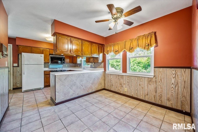 kitchen with white refrigerator, light tile patterned flooring, wood walls, kitchen peninsula, and ceiling fan