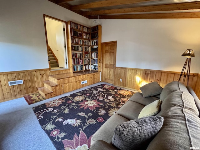 carpeted living room with beam ceiling, wainscoting, wood walls, and stairway