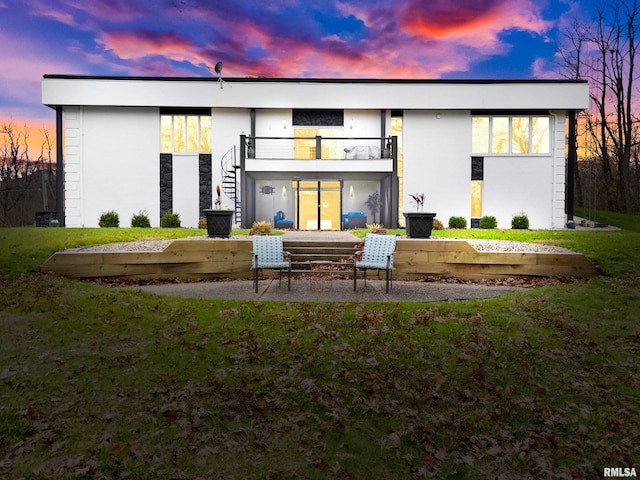 back house at dusk featuring a balcony, a patio area, and a lawn