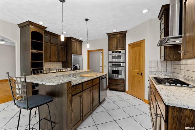 kitchen featuring wall chimney exhaust hood, stainless steel appliances, a kitchen island with sink, sink, and pendant lighting