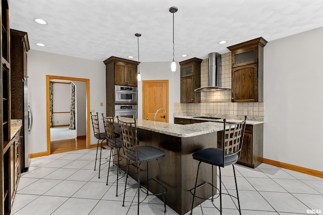 kitchen with wall chimney range hood, backsplash, an island with sink, decorative light fixtures, and light tile patterned floors