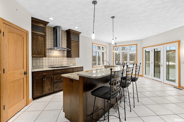 kitchen with light stone countertops, wall chimney exhaust hood, black electric cooktop, a kitchen island with sink, and pendant lighting