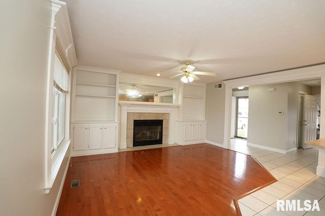 unfurnished living room with built in shelves, ceiling fan, light wood-type flooring, and a tiled fireplace