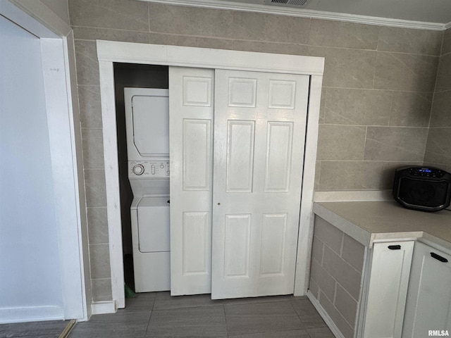 washroom featuring dark tile patterned floors, stacked washing maching and dryer, tile walls, and ornamental molding