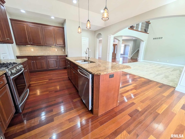 kitchen with appliances with stainless steel finishes, dark wood-type flooring, sink, a center island with sink, and decorative light fixtures