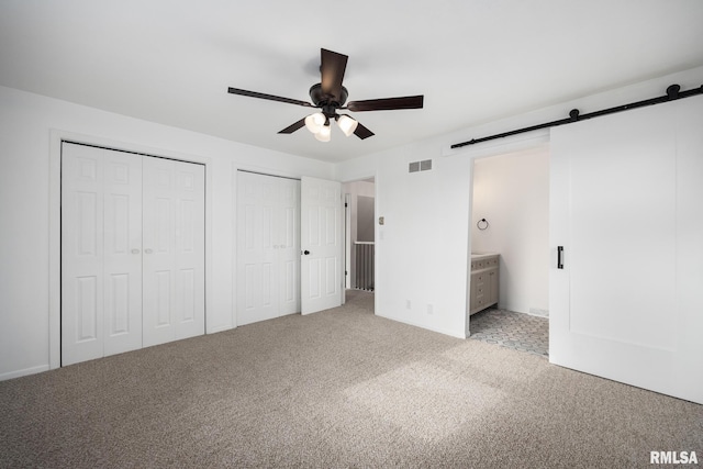 unfurnished bedroom featuring ensuite bath, light colored carpet, ceiling fan, a barn door, and multiple closets