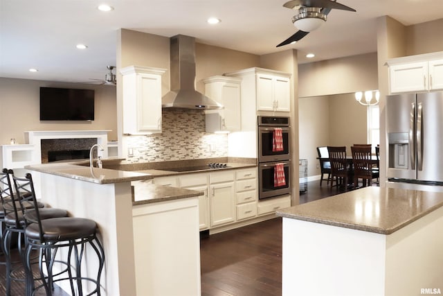 kitchen featuring a kitchen breakfast bar, wall chimney range hood, ceiling fan, white cabinetry, and stainless steel appliances