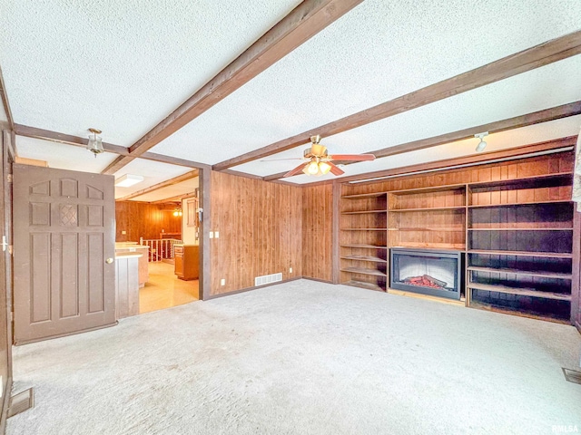 unfurnished living room with a textured ceiling, beamed ceiling, light carpet, ceiling fan, and wood walls