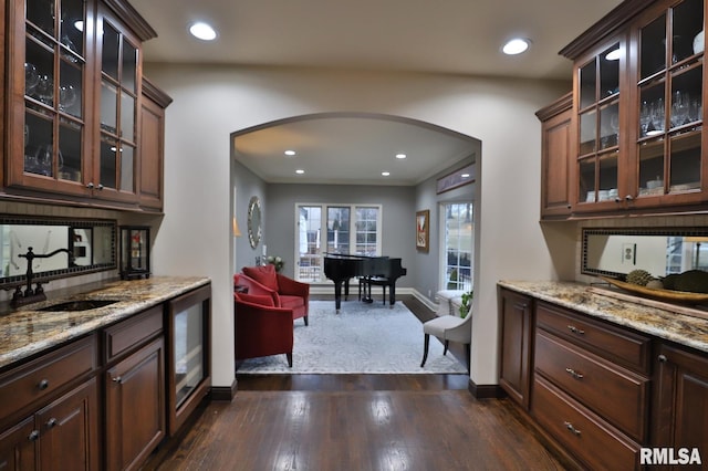 bar featuring sink, decorative backsplash, dark brown cabinetry, light stone countertops, and dark wood-type flooring