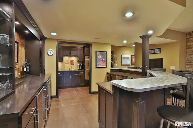 kitchen with ornate columns, a kitchen breakfast bar, a kitchen island with sink, light tile patterned floors, and dark brown cabinetry