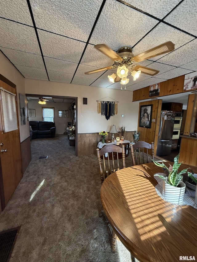 carpeted dining room with ceiling fan, a paneled ceiling, and wood walls