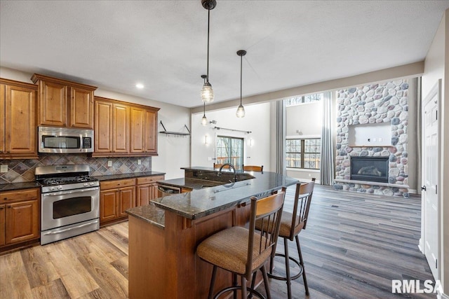 kitchen with a stone fireplace, a center island with sink, appliances with stainless steel finishes, pendant lighting, and backsplash