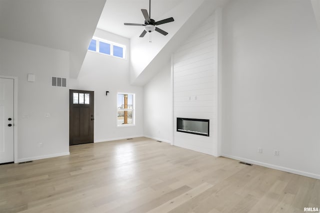 unfurnished living room featuring ceiling fan, a large fireplace, light wood-type flooring, and a towering ceiling