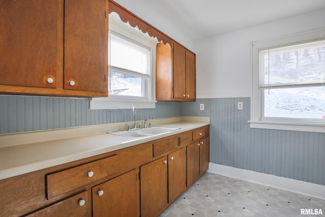 kitchen with brown cabinetry, a wainscoted wall, light countertops, light floors, and a sink