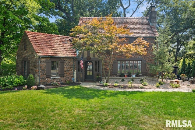 english style home featuring a tile roof, brick siding, a chimney, and a front lawn