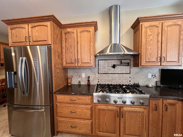 kitchen featuring appliances with stainless steel finishes, brown cabinets, dark stone countertops, wall chimney range hood, and backsplash