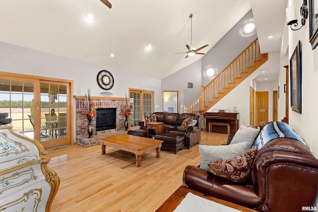 living area featuring light wood finished floors, ceiling fan, stairs, a brick fireplace, and high vaulted ceiling