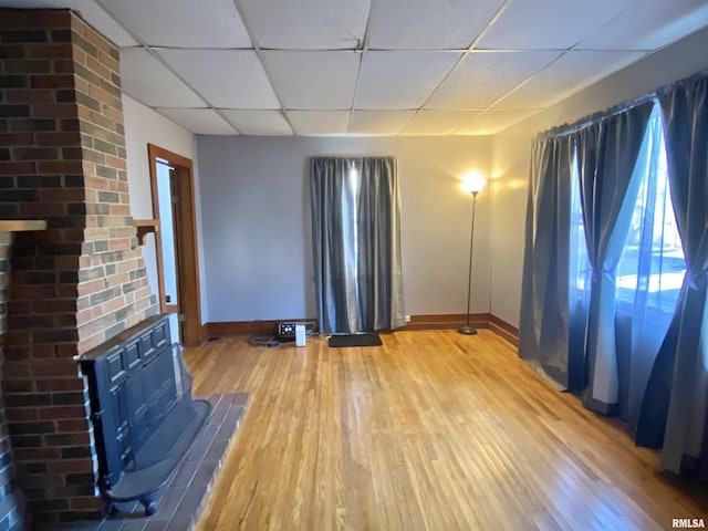 unfurnished living room featuring a wood stove, a drop ceiling, light wood-style flooring, and baseboards