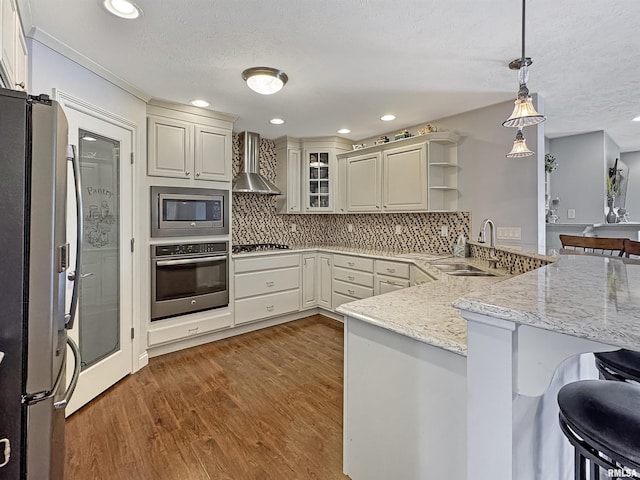 kitchen featuring a peninsula, a kitchen breakfast bar, stainless steel appliances, wall chimney exhaust hood, and a sink