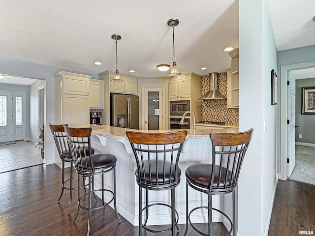 kitchen featuring a peninsula, cream cabinetry, appliances with stainless steel finishes, wall chimney exhaust hood, and tasteful backsplash