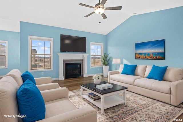 living area with baseboards, a glass covered fireplace, lofted ceiling, ceiling fan, and wood finished floors