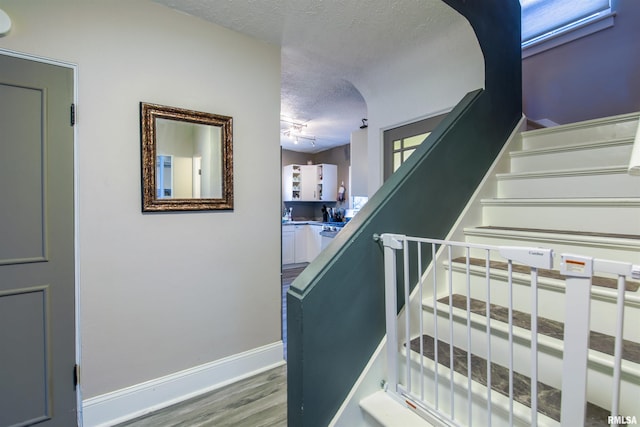 stairway featuring rail lighting, baseboards, a textured ceiling, and wood finished floors