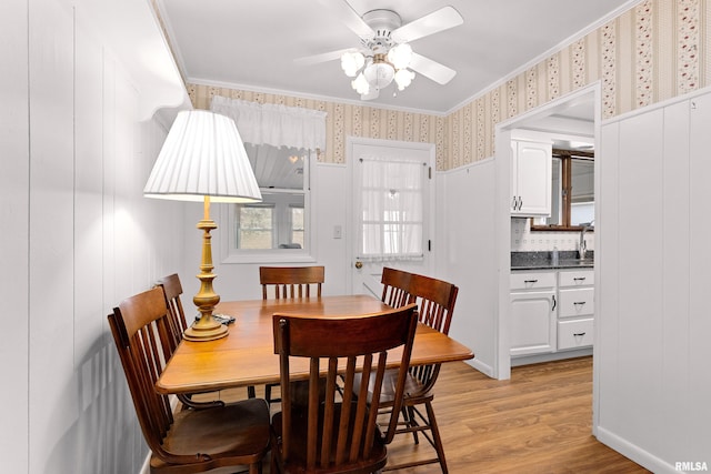 dining room featuring light wood finished floors, ornamental molding, a ceiling fan, and wallpapered walls