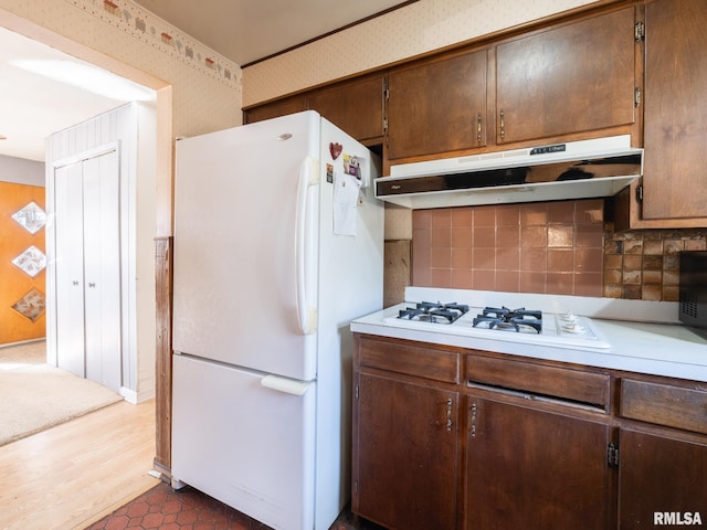 kitchen with under cabinet range hood, wallpapered walls, white appliances, dark brown cabinetry, and light countertops