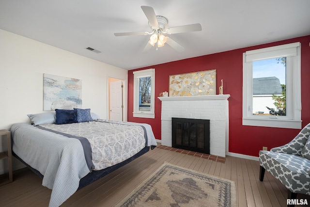 bedroom featuring baseboards, visible vents, ceiling fan, wood-type flooring, and a brick fireplace