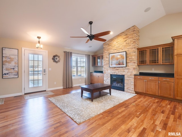 living room with baseboards, ceiling fan, light wood-type flooring, a stone fireplace, and high vaulted ceiling