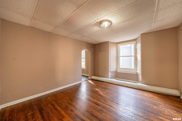 unfurnished room with hardwood / wood-style floors, arched walkways, baseboards, and a drop ceiling