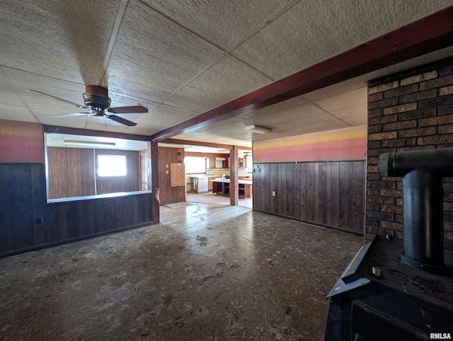 unfurnished living room with a ceiling fan, a paneled ceiling, wooden walls, and a wood stove