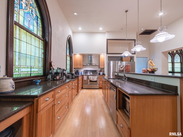 kitchen with a towering ceiling, stainless steel appliances, dark stone countertops, a sink, and wall chimney range hood