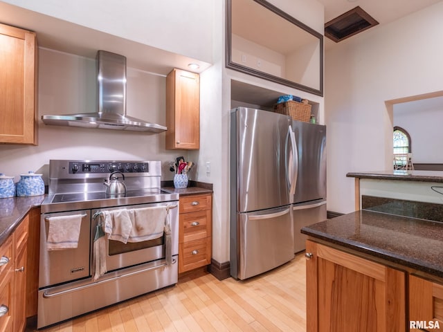 kitchen featuring stainless steel appliances, dark stone counters, light wood-style floors, wall chimney range hood, and brown cabinetry