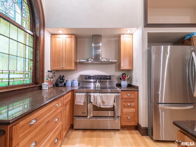 kitchen featuring wall chimney range hood, stainless steel appliances, dark stone counters, and light wood finished floors
